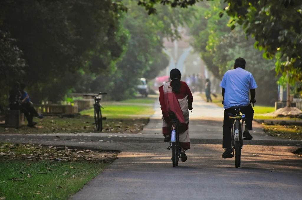 The image shows a man and a woman riding bicycles down a broad concrete path. There is grass growing on either side of the path and there are lush green trees lining growing behind the grass patches. There are people sitting on a bench on the side._Urban planning
