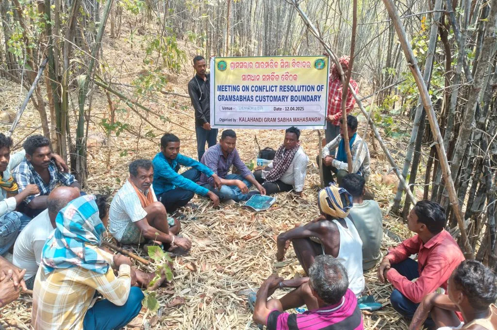 a group of men seated on the floor of a wooded area, with a banner saying "Meeting on conflict resolution of Gram Sabhas customary boundary" in the back--commons governance